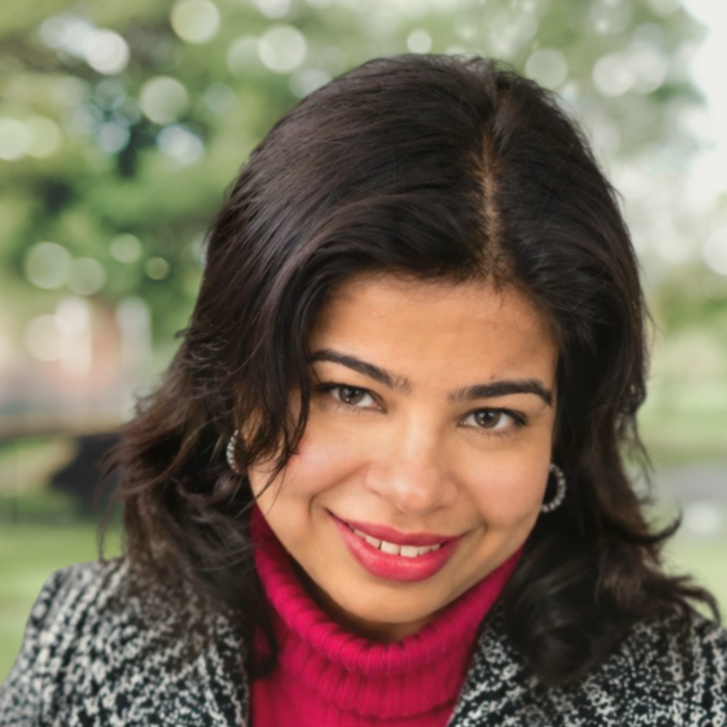 Close-up portrait of a smiling woman with dark hair wearing a red knit turtleneck and a black-and-white patterned coat, outdoors with blurred green background.